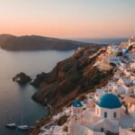 Santorini budget travel tips aerial view of caldera with white buildings and blue domes at sunset